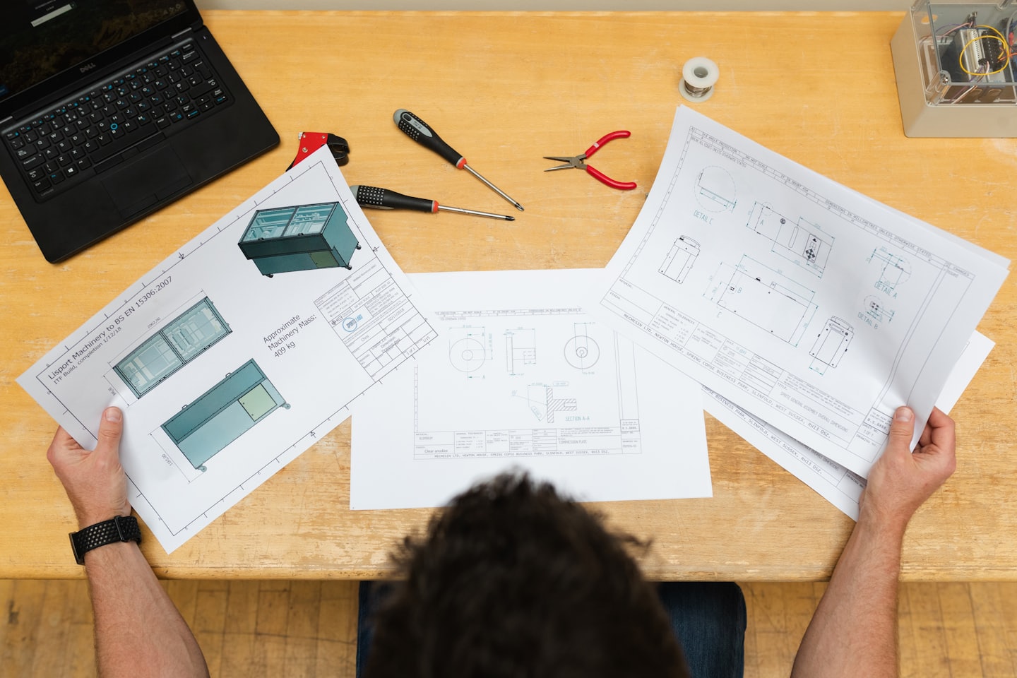 Person reviewing blueprints with tools and laptop on desk.
